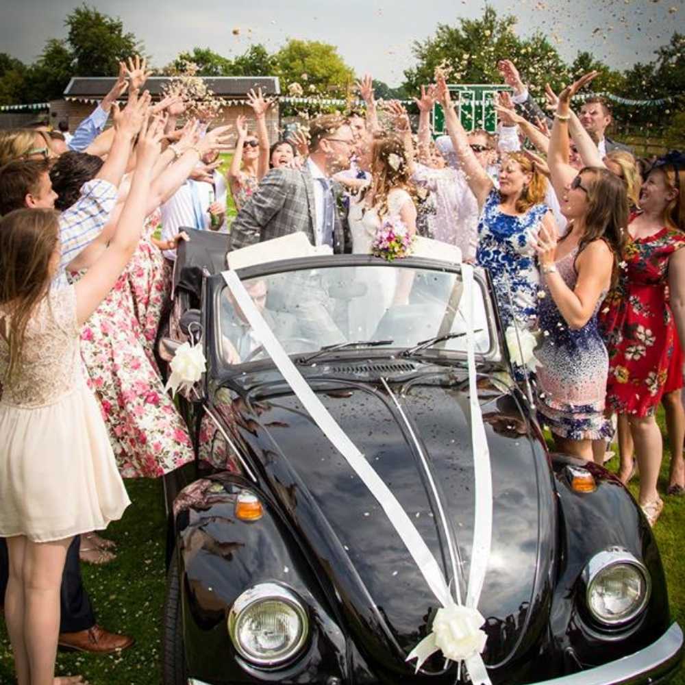A bride and groom sharing Confetti Kisses in a Beetle car! They are surrounded by their wedding guests throwing real flower petal confetti.